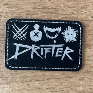 May include: A rectangular black patch with white stitching around the edges. The patch features the word DRIFTER in a stylized font, with four icons above it: three diagonal slashes, a silhouette with an X, a set of fangs, and a splatter shape.