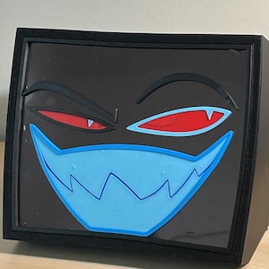 May include: A black box with a dark screen featuring a stylized face. The face has two narrow red eyes with black eyebrows and a wide, light blue mouth with sharp, jagged teeth. The entire design is layered and raised against the dark background.