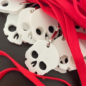 May include: White skull-shaped pendants with large eye and nose holes are attached to red lanyards. The pendants are made of a smooth material and have a simple, clean design. The red lanyards are thick and woven.