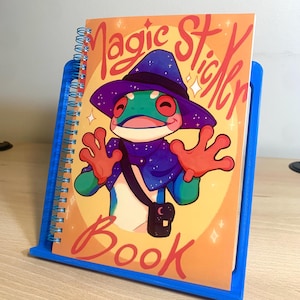 Puede incluir: Un cuaderno de espiral azul con una portada colorida que presenta una rana de dibujos animados con un sombrero de mago morado y azul. La portada dice "Magic Sticker Book".