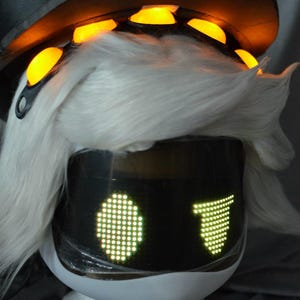 May include: A white wig topped with a black cap featuring glowing orange circular accents. The front displays a dark visor with two illuminated green LED pixelated eyes, one oval and one triangular, set on a white base structure.
