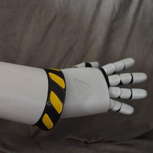 May include: A white robotic arm featuring a smooth, cylindrical forearm and a multi-jointed hand. A black bracelet with diagonal yellow hazard stripes is worn around the wrist area, providing a high-contrast detail against the white surface.