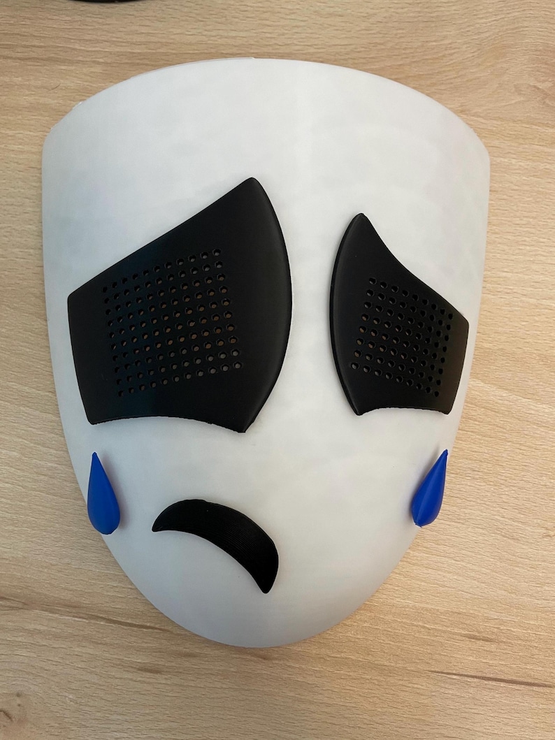 May include: A white mask with a sad expression. The mask has black eyes with a grid pattern, a black frown, and two blue teardrop shapes.