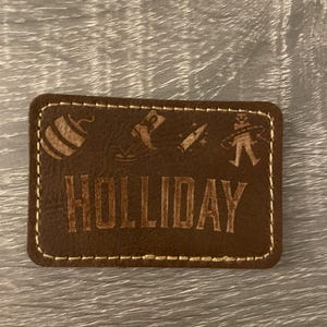 May include: A rectangular brown leather patch with light-coloured stitching around the edges. The word HOLLIDAY is engraved in the centre. Above the text are small engraved icons of a barrel, a cowboy boot, a bullet, and a person with a hula hoop.