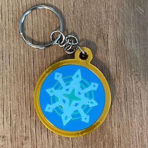 May include: A round keychain with a gold border and a blue center featuring a snowflake design in shades of light blue and green. The keychain has a silver ring for attaching keys.