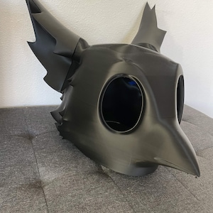 以下が含まれることがあります： マットブラックの立体的な動物型マスク。ギザギザした縁を持つ大きな尖った耳、突き出たクチバシのような鼻先、そして2つの大きな空洞の目穴が特徴です。滑らかな質感で、はっきりとした鋭い輪郭に成形されています。