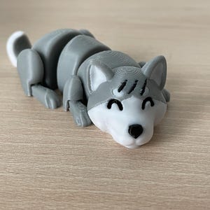 Könnte beinhalten: Ein graues und weißes, 3D-gedrucktes, artikuliertes Spielzeug-Hündchen mit einem fröhlichen Ausdruck. Der Hund liegt mit dem Kopf auf der Oberfläche.