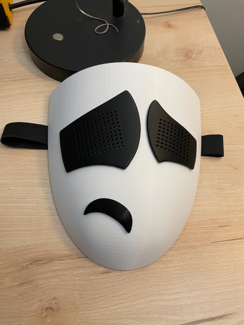 May include: A white plastic mask with black eye holes and a frown mouth. The mask has a black elastic band for wearing.