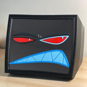 Puede incluir: Caja negra con un diseño de cara de dibujos animados. La cara tiene ojos rojos, una boca azul con dientes blancos y un fondo negro. La caja está sobre una superficie marrón claro.