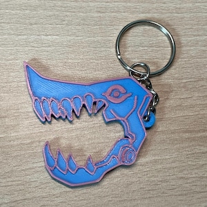 May include: A blue and pink 3D printed keychain shaped like a skull with a large open mouth and sharp teeth. The keychain has a silver metal ring attached to it.