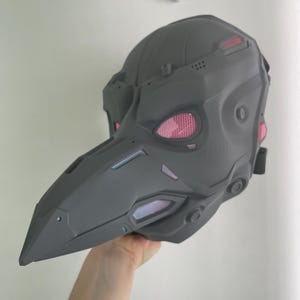 May include: A matte dark gray plague doctor mask with a long, pointed beak and angular, mechanical-style paneling. The eye sockets feature pink mesh lenses, and there are small pink accents along the sides and beak, with a black strap visible.