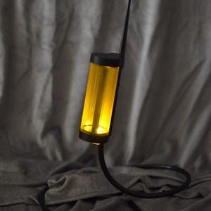 May include: A cylindrical yellow light fixture with a black top and base, featuring a thin black antenna-like protrusion extending upward. The light is connected to a long, flexible black cord that curves along the surface below the unit.