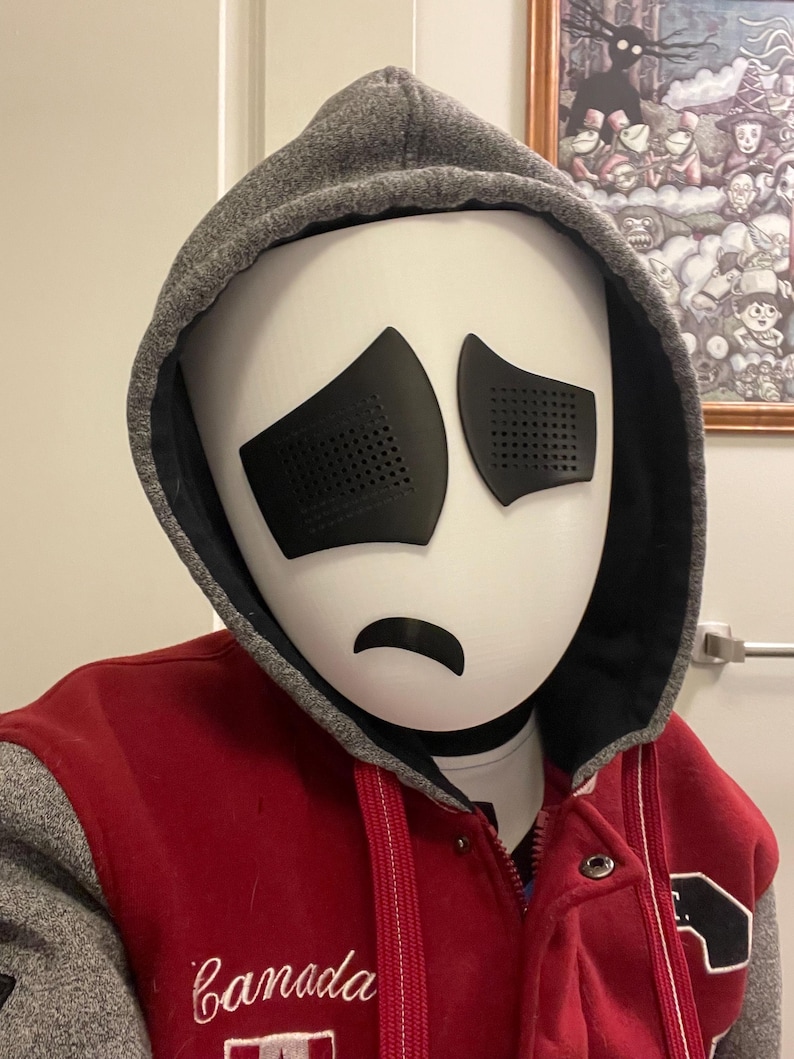 May include: A white mask with black eyes and a frown. The mask is worn by a person wearing a red and grey hooded sweatshirt with the word "Canada" embroidered on the front.