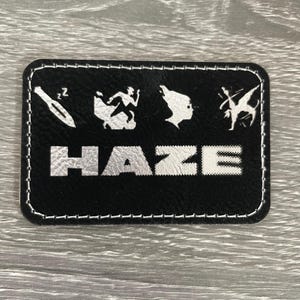 May include: A rectangular black patch with rounded corners and white stitched edges. It features the word HAZE in large white letters, topped by four white icons: a sleeping knife, a running figure, a profile silhouette, and a gymnast.