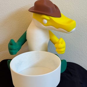 Puede incluir: Una caprichosa y colorida figura de un caimán con un sombrero de vaquero marrón. El caimán tiene la cabeza amarilla, el cuerpo blanco y los brazos y las piernas verdes. Un cuenco blanco está delante de la figura.