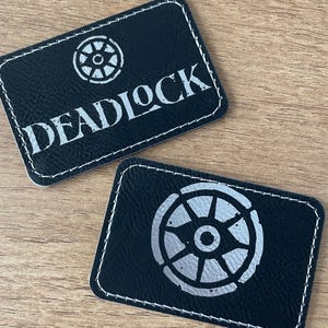 May include: Two rectangular black patches with white stitched borders. One patch features a silver wheel icon above the text DEADLOCK. The other patch displays a large silver wheel icon. Both patches have a textured surface and rounded corners.