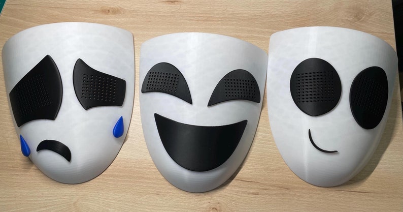 May include: Three white masks with different facial expressions. The first mask has a sad expression with black eyes and a frown, the second mask has a happy expression with black eyes and a smile, and the third mask has a neutral expression with black eyes and a small smile.