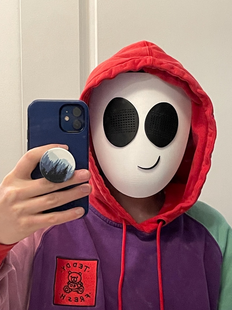 May include: A person wearing a red and purple hoodie with a white mask featuring black eyes and a smile. The hoodie has a red patch with a cartoon bear and the text "FRE$H TEDDY".