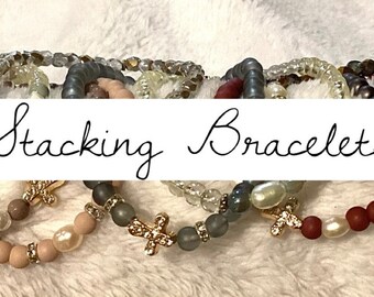 May include: A set of stacking bracelets featuring various colors and textures, including brown, beige, gray, and red. The bracelets are made of beads and have a variety of charms, including crosses and pearls. The text "Stacking Bracelets" is written in black cursive on a white background.