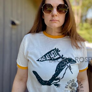 May include: A white t-shirt with a yellow collar and cuffs featuring a black illustration of a lizard holding a flag that reads "MAD AS HELL." The shirt also has the text "BLOOD ROSE DESIGN CO." and the year "2023."