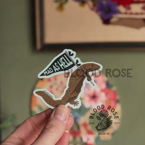 May include: A sticker featuring a brown salamander holding a flag that reads "MAD AS HELL." The sticker has a white border and the words "BLOOD ROSE" are printed on the salamander's body. The sticker is held by a person's hand.
