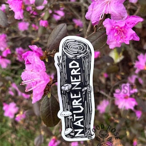 May include: A black and white sticker with the text "NATURE NERD" and mushroom illustrations. The sticker is set against a backdrop of pink flowers and green foliage. The sticker also includes the text "BLOOD ROSE" and "DESIGN CO."