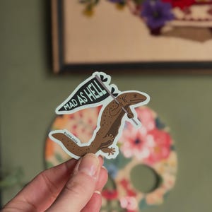 May include: A sticker featuring a brown salamander holding a flag that reads "MAD AS HELL." The sticker has a white border and the text "BLOOD ROSE" is printed on the bottom left. The background is a floral skull.
