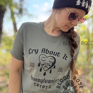 May include: Sage green t-shirt with the text "Cry About It" and a sad heart graphic. The shirt also features the words "Pennsylvania's Cryptid" and the Blood Rose logo. A black beanie with white ghost figures and heart-shaped sunglasses complete the look.