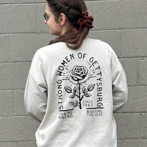 May include: Cream-colored sweatshirt with a black graphic on the back. The graphic features a rose and the text "STRONG WOMEN OF GETTYSBURG SINCE 1863 BAKING BREAD BURYING THE DEAD."