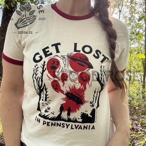 May include: Cream-colored t-shirt with a maroon trim featuring a graphic design. The design includes the text "GET LOST IN PENNSYLVANIA" with illustrations of a UFO, ghosts, and a car. The shirt has a vintage aesthetic.