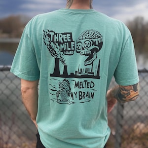 May include: Seafoam green t-shirt with a black graphic design. The design includes the text "THREE MILE ISLAND" above a depiction of smokestacks, a skull with a melting brain, and the text "MELTED MY BRAIN".