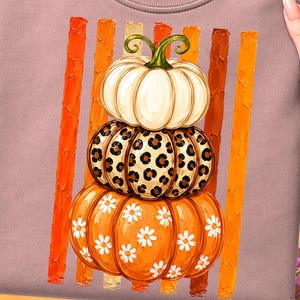 May include: A mauve crewneck t-shirt featuring a graphic of three stacked pumpkins. The top pumpkin is cream, the middle has a brown leopard print, and the bottom is orange with white daisies. Vertical orange and brown brushstroke stripes appear behind.