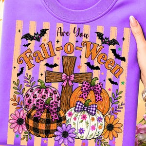 May include: A purple crewneck sweatshirt featuring a graphic print with the text Are You Fall-o-Ween. The design includes a wooden cross, pumpkins with leopard, plaid, and floral patterns, bows, bats, and flowers against a striped background.