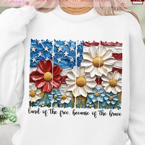 Può includere: Una felpa bianca con una grafica di una bandiera americana fatta di fiori. Il testo "land of the free. because of the brave" è stampato sotto la grafica.