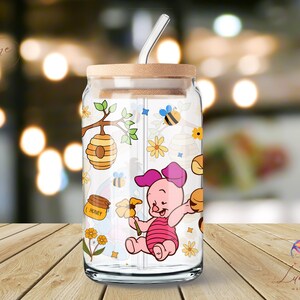 May include: A clear glass jar with a straw and a wooden lid. The jar has a colorful design featuring a cartoon pig, honeycombs, bees, and flowers. The text "Honey" is printed on the jar.