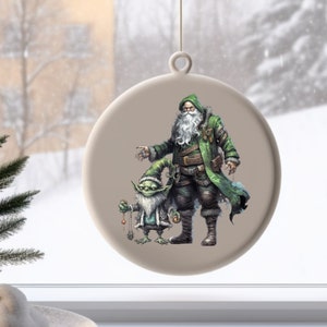 Op de afbeelding: Een ronde, witte kerstbal met een groen geklede kerstman en een groene kobold die een reeks ornamenten vasthoudt. De kerstbal is versierd met een beige achtergrond.