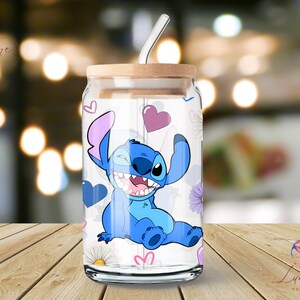 Op de afbeelding: Een transparante glazen beker met een blauw en paars cartoonfiguur erop. Het figuur is Stitch uit de Disney-film Lilo & Stitch. De beker heeft een rietje en een houten deksel. De achtergrond is wazig en onscherp.