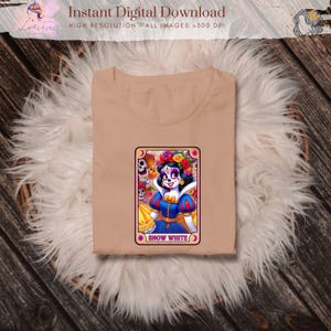 Może przedstawiać: Beżowa koszulka z kolorową grafiką Kr&oacute;lewny Śnieżki w stylu Dnia Zmarłych. Grafika znajduje się na karcie z napisem "Snow White". Koszulka jest złożona na puszystej, białej powierzchni.