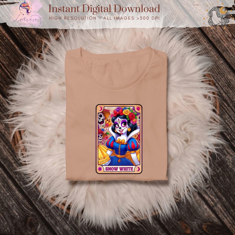 Może przedstawiać: Beżowa koszulka z kolorową grafiką Kr&oacute;lewny Śnieżki w stylu Dnia Zmarłych. Grafika znajduje się na karcie z napisem "Snow White". Koszulka jest złożona na puszystej, białej powierzchni.