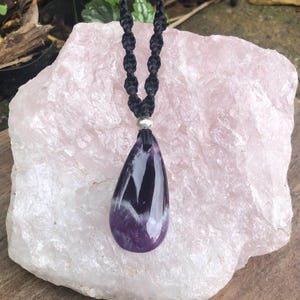 May include: A purple amethyst pendant necklace with a black woven cord. The teardrop-shaped stone has white and dark purple veining. A small silver bead joins the pendant to the cord. The necklace is shown on a large, light pink stone.