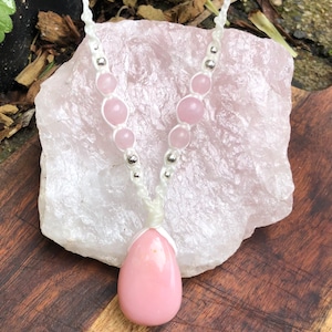May include: A pink gemstone pendant necklace with a white cord and silver beads. The necklace is displayed on a large pink crystal.