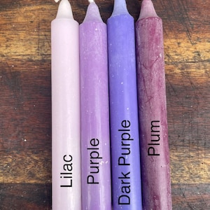 Może przedstawiać: Cztery cylindryczne świece stożkowe ustawione obok siebie w gradiencie odcieni fioletu. Świece są oznaczone napisami Lilac, Purple, Dark Purple oraz Plum, odpowiadającymi ich kolorom od najjaśniejszego do najciemniejszego.