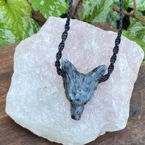 May include: A handcrafted necklace featuring a carved stone pendant in the shape of a wolf's head. The pendant is gray with black accents and is suspended from a black braided cord. The necklace is displayed on a light pink stone.