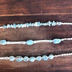 May include: Three handmade necklaces with white macramé cords and light blue beads. The top necklace features small, irregular beads, while the others have larger, oval-shaped beads. The necklaces are displayed on a dark wooden surface.