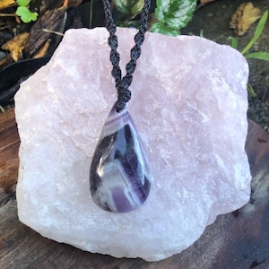 May include: A teardrop-shaped amethyst pendant necklace with white and purple stripes, suspended from a black woven cord. The gemstone sits on a large, light pink crystal. The pendant is a beautiful piece of jewellery.