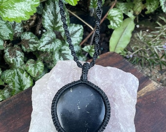 May include: A round, smooth black stone pendant encased in a black macrame cord setting. The pendant hangs from a matching black knotted cord necklace, featuring a circular frame crafted from intricate, tightly woven dark fiber thread work.