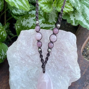 May include: A necklace featuring a teardrop-shaped rose quartz pendant suspended from a dark brown macrame cord. The cord is accented with smaller round rose quartz beads and metallic silver-toned beads arranged symmetrically on both sides.