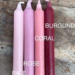 Może przedstawiać: Cztery cylindryczne świece ustawione pionowo, prezentujące gradient odcieni r&oacute;żu i czerwieni. Kolory są oznaczone jako PALE PINK, ROSE, CORAL i BURGUND. Każda świeca ma gładkie, matowe wykończenie woskowe oraz mały, niezapalony bawełniany knot.