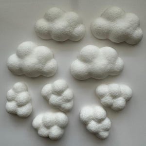 Puede incluir: Nueve objetos decorativos blancos con forma de nube. Cada nube tiene una apariencia texturizada y esponjosa, con diferentes tamaños y formas. Las nubes están dispuestas sobre un fondo blanco liso, creando una estética caprichosa.
