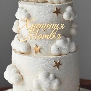 May include: A two-tiered white cake with a textured finish, decorated with white fondant clouds and metallic gold star accents. A gold script cake topper with the Cyrillic text "Хрещення Матвія" is positioned on the upper tier of the cake.